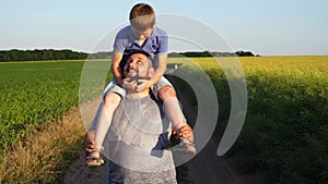 Daddy goes with small son sitting on his shoulders through rural road near green meadows. Happy father and his little