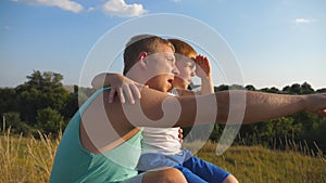 Daddy pointing at something to his little kid. Dad and child sitting at green grass on the hill and speaking with each