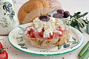 Dakos traditional greek appetizer on a traditional plate with ceramic olive oil jar, dry rye bread, olives and olive branch.