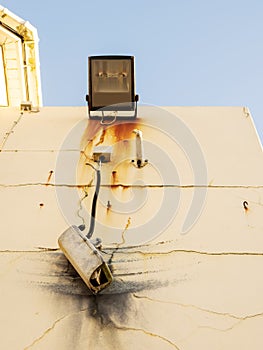 Damaged CCTV camera hanging on a cable on yellow wall, old flood light above the camera. Front element is broken and there is sign