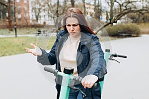 Damaged e-scooter for rent. Beautiful redhead 30s woman standing on modern scooter, frowning upset because of problem