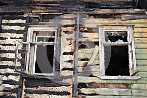 The damaged wall of the house from the fire and two windows with broken glass in the rays of the setting sun in the spring