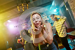 Dance and Party. Attractive caucasian young woman blowing confetti to camera while posing in the night club. Friends