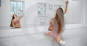 Dancer is sitting on the floor and warming up in front of the mirror wall in the white bright dance hall, ballet