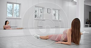 Dancer is sitting on the twine and warming up infront of the mirror wall in the white bright dance hall, ballet