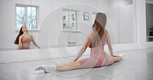 Dancer is sitting on the twine and warming up infront of the mirror wall in the white bright dance hall, ballet