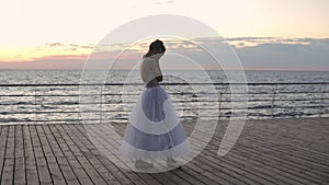 Dancing ballerina exercising on embankment above ocean or sea at sunrise or sunset. Young woman a bit upset about