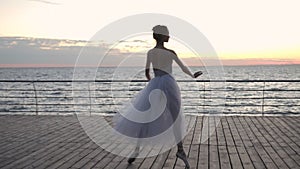 Dancing ballerina in white ballet tutu and pointe on embankment above ocean or sea at sunrise or sunset. Young