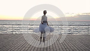 Dancing ballerina in white tutu and pointe on embankment above ocean or sea beach at sunrise. Young beautiful woman