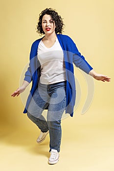 Dancing beautiful young woman plus size in jeans and a blue jacket. Yellow background. Vertical