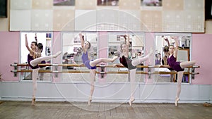 In dancing hall, Young ballerinas in purple leotards perform developpe attitude efface on pointe shoes, raise their legs