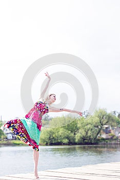Dancing joy: beautiful blond young slim woman enjoying stretching in long light dress at water lake on summer green outdoors