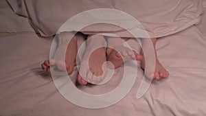 Dancing legs of boy son child and mother female foot lie under the blanket on sheet in the evening. Happy baby kid girl