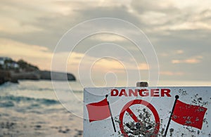 Danger, no swimming sign with red flags, blurred sea background with cliff shore, morning overcast sky above