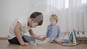 Danger to children, boy teaches little brother to turn on and off the iron in electric socket sitting on the floor in