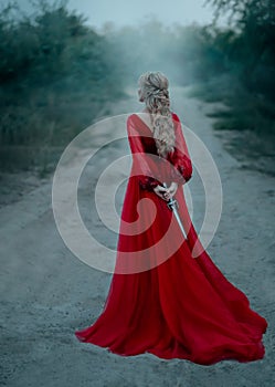 Dangerous blonde queen in red fashion lush dress Hides a dagger behind. Backdrop dark fantasy forest in fog. Concept