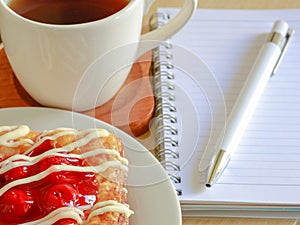 Danish pastry with a cup of hot tea and a pen and small notebook on wood table in morning time and red heart for valentine