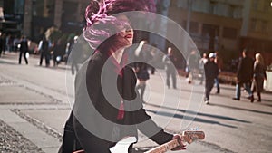 Daring rebel woman with purple hair playing electric guitar at city center during bright sunset. People walking in