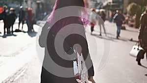 Daring rebel woman with purple hair walking with electric guitar at city center during bright sunset. People walking in