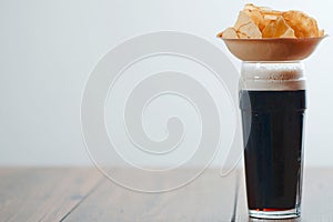 Dark ale in a glass with foam and a wooden small cup with potato chips on a wooden table. Selective focus. Traditional beer and