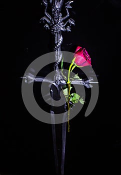 dark background with a red rose with a green stem resting on a black surface flanked by a sword