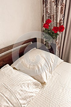 A dark bed with white and lilac bedding stands next to a bedside table on which there is a glass vase with a bouquet of roses