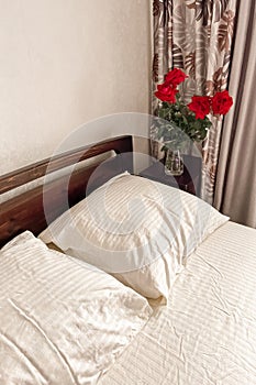 A dark bed with white and lilac bedding stands next to a bedside table on which there is a glass vase with a bouquet of roses.