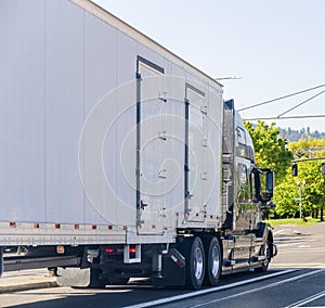 Dark big rig long haul semi truck transporting commercial cargo in covered semi trailer driving on the city street