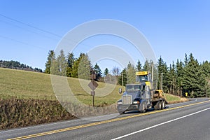 Dark big rig semi truck transporting tractor on flat bed semi trailer running on the road with field on the hill