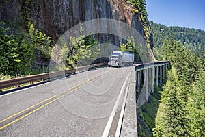 Dark blue big rig day cab semi truck transporting cargo in bulk semi trailer driving on the mountain road with rock wall and