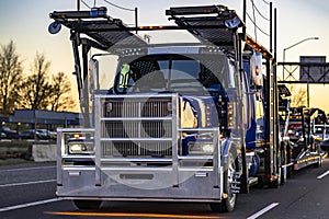 Dark blue classic big rig car hauler semi truck transporting cars on modular semi trailer running on the highway road at twilight