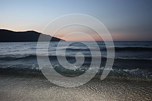 Dark blue sea waves over light sand with mountain and sunset on horizon