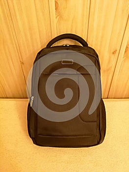 Dark brown backpack, with handle and zippers, leaning against wooden paneling