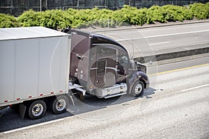 Dark brown big rig commercial long haul semi truck transporting dry van semi trailer on the divided highway road