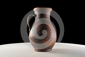 Dark brown jug with a handle. Handmade jar from clay standing on white table isolated on black studio background