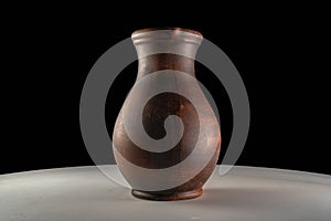 Dark brown jug. Handmade jar from clay standing on white table isolated on black studio background. Earthenware for