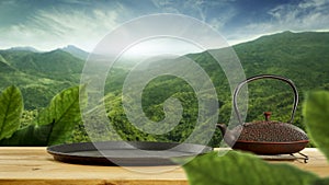 A dark brown teapot and black tray on wooden table with beautiful green trees mountains and blue cloudy sky background.