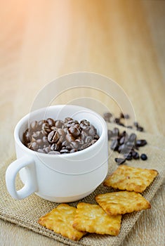 Dark coffee beans in coffee mug with cracker on wood table.