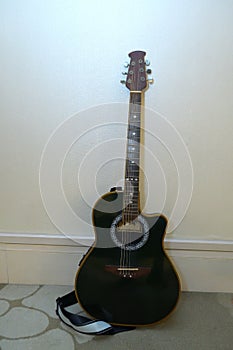 A dark colored acoustic guitar with shoulder strap resting upright against a white wall