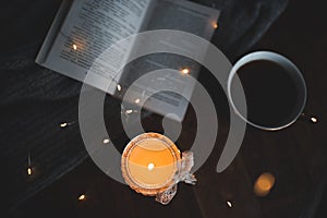 Dark and cozy atmosphere in room: open paper book with burning candle and cup of fresh coffee on wooden table closeup top view at