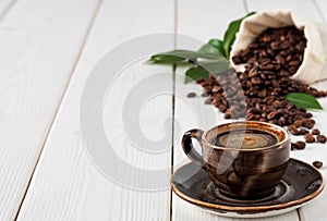 Dark cup coffee espresso on a saucer and coffee beans in burlap sack on a white wooden background. Close up, soft focus, copy