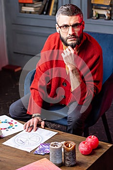 Dark-eyed astrologist wearing red cardigan sitting at the table with candles