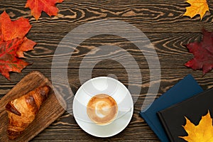Dark flat lay with a cup of coffee, homemade croissant, books and colorful maple leaves on the wooden background.