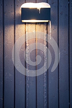 Dark, frozen house wall with icicles and street light background. Old, grey, vertical wooden boards with shine. Winter holidays