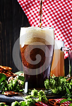 Dark German beer is poured into a glass, fresh green hops and bo