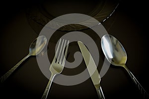 Dark glass plate, silver knife, fork, spoon, teaspoon on the dark reflective backdrop.