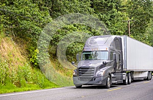 Dark gray big rig semi truck with dry van semi trailer delivering cargo running on the mountain forest green road