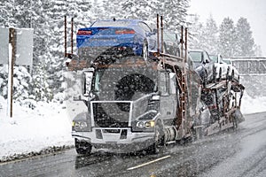 Dark gray car hauler big rig semi truck tractor transporting cars on modular semi trailer driving slowly and carefully on a snowy