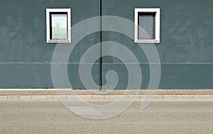 Dark gray plaster wall with gutter in the middle and two windows by sides. Sidewalk and street in front