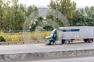 Dark green big rig day cab semi truck transporting cargo in short semi trailer driving on the divider road